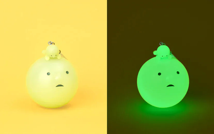 A figurine of a simplistic cartoonish head, frowning slightly, while looking up at a second smaller figure laying on its head. A second image shows the same figurine glowing in the dark.