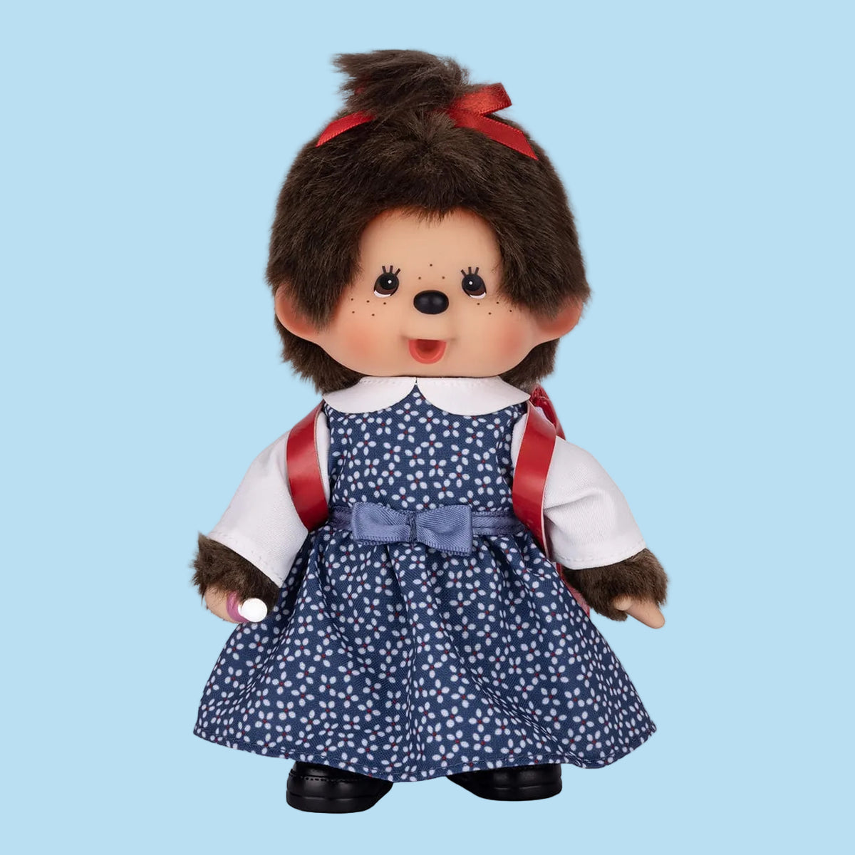 A Monchhichi plush wearing a blue dress with white polka dots, and a red backpack. 