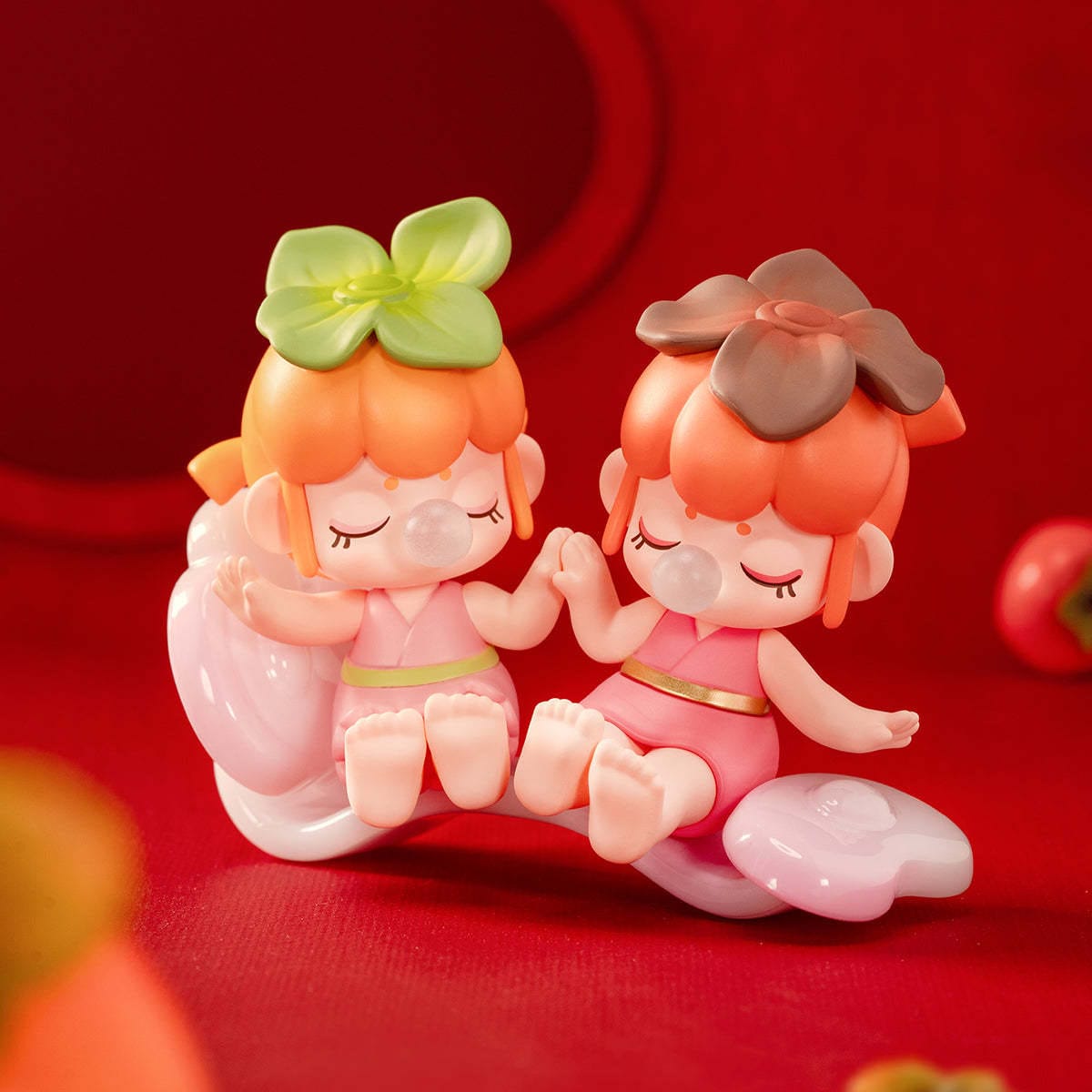 A plastic figure of two little girls sitting together. They're both blowing bubbles of gum and wearing leaves on their head.