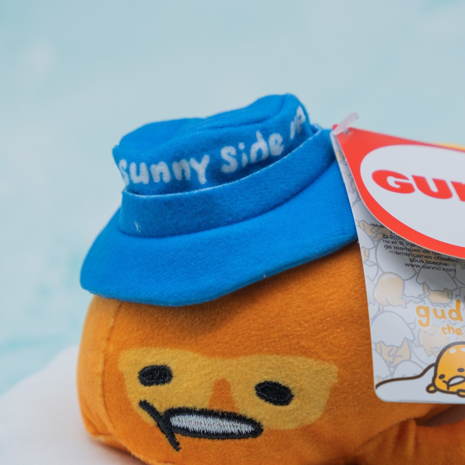 Plush of an egg yolk with a lazy expression wearing a blue hat that says "sunny side up".