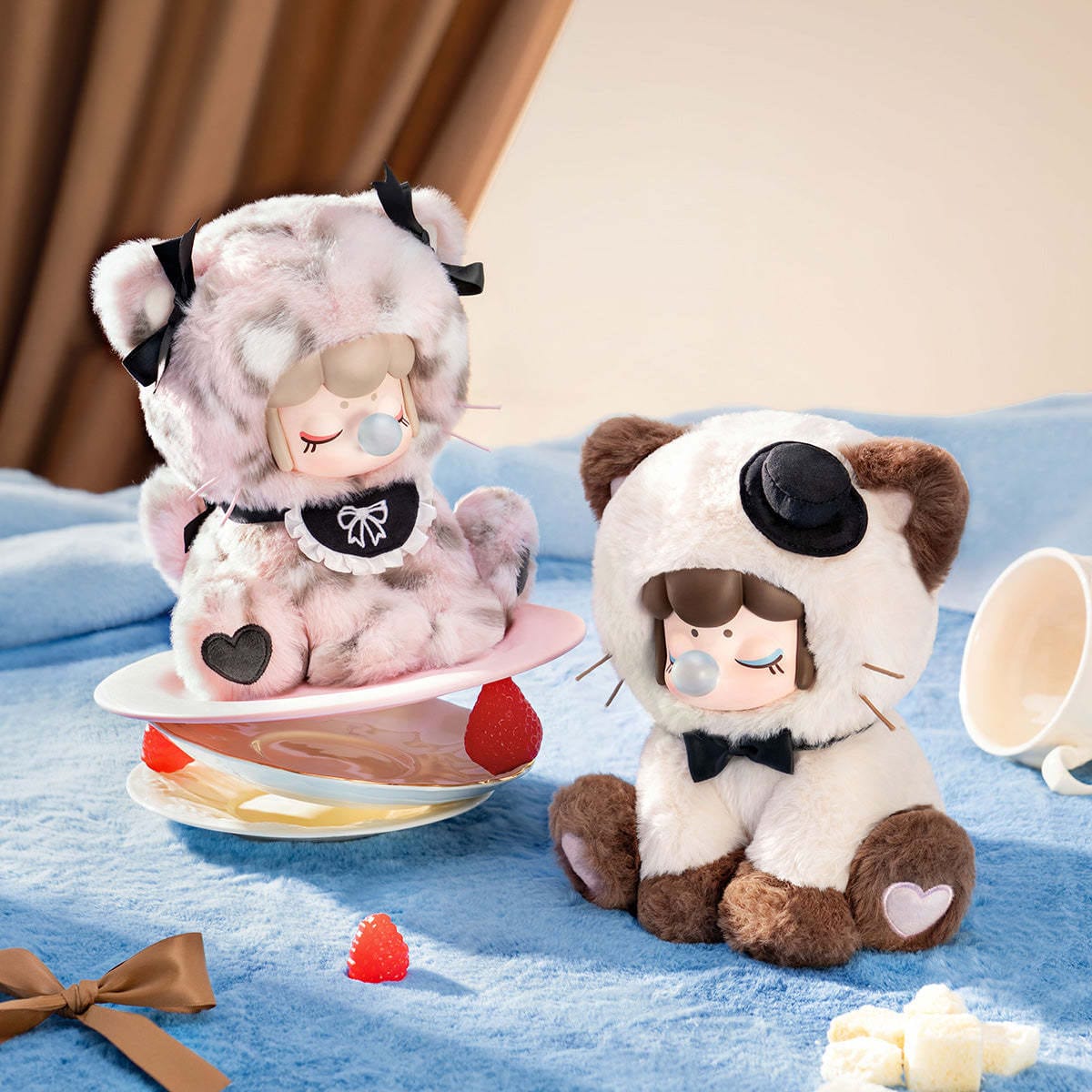 Two figures of little girls in fluffy cat onesies. One has a little hat and a bow tie, while the other has an apron and ribbons.