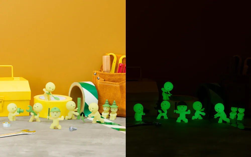 Two seperate images beside each other. The first image is of many different construction related Smiskis, with some holding tools and others wearing traffic cones as hats. The second image is a copy of the first but in darkness showing the Smiskis glowing.